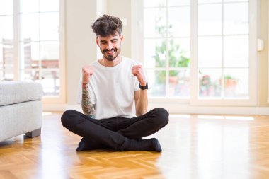 Genç adam evde rahat rahat oturup mutlu ve heyecanlı bir şekilde kolları havada, gülümseyerek ve başarı için çığlık atarak zafer jesti yapıyor. Kutlama konsepti.