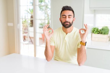 Yakışıklı, İspanyol asıllı sıradan sarı tişörtlü evde rahatla ve gözleri kapalı gülümse parmaklarınla meditasyon hareketi yap. Yoga konsepti.