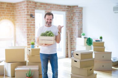 Orta yaşlı bir adam yeni bir eve taşınıyor. Karton kutular paketliyor. Başparmağıyla işaret ediyor ve gülümsüyor.