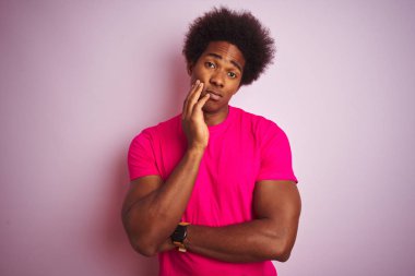 Siyah saçlı genç Amerikalı adam pembe arka planda dikilip kollarını kavuşturup depresyondan sıkılmış ve yorgun göründüğünü düşünüyor..