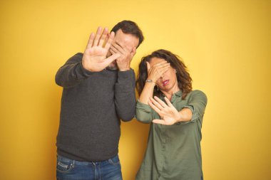 Güzel bir orta yaşlı çift, izole edilmiş sarı arka planda, elleriyle gözleri kapatıyor ve üzgün ve korku ifadeleriyle jest yapıyor. Utanç verici ve olumsuz bir kavram..