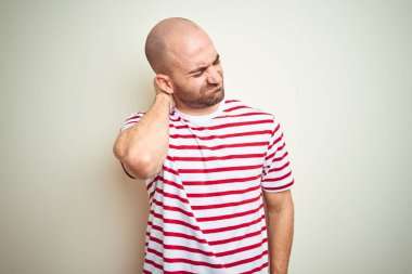 Sakallı genç ve kel bir adam, beyaz izole edilmiş arka planda günlük kırmızı tişört giyiyor boynu ağrıyor, boynuna elle dokunuyor, kasları ağrıyor.