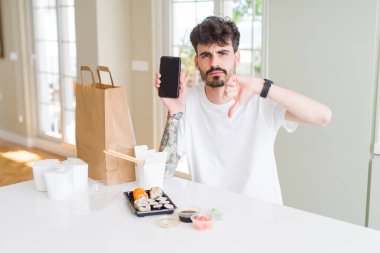 Eve servis yaparken Asya suşisi yiyen ve kızgın yüzlü akıllı telefon ekranı gösteren genç bir adam. Negatif işaretler, baş parmakları aşağıda, reddedilme konsepti.