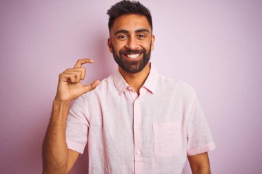 İzole edilmiş pembe arka planda dikilen rahat gömlekli genç Hintli adam gülümsüyor ve el hareketleriyle küçük beden işareti yaparak kameraya bakıyor. Ölçü kavramı.