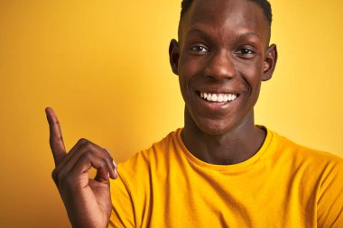 Siyah tişörtlü Afro-Amerikan bir adam izole edilmiş sarı arka planda dikilirken mutlu bir şekilde elini ve parmağını kenarı gösteriyor.