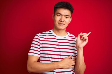 Asyalı genç bir Çinli, çizgili tişört giyiyor. İzole edilmiş kırmızı arka planda duruyor. Yüzünde kocaman bir gülümseme var..