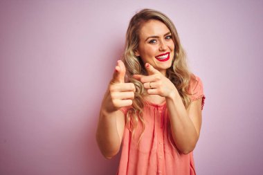 Pembe izole edilmiş arka planda tişört giyen genç ve güzel bir kadın mutlu ve komik yüzlü parmaklarını kameraya doğrultuyor. İyi enerji ve titreşimler.