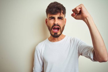 İzole edilmiş beyaz arka plan üzerinde duran dövmeli genç adam öfkeyle bağırırken kızgın ve öfkeli bir şekilde yumruk kaldırıyor. Öfke ve agresif kavram.