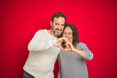 Güzel bir orta yaşlı çift, izole edilmiş kırmızı arka plan üzerine kazak giyip, ellerinde kalp sembolü olan elleri ile gülümsüyorlar. Romantik konsept.