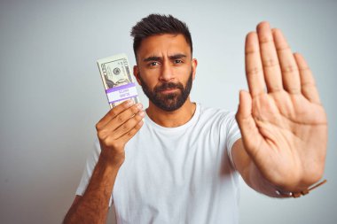İzole edilmiş beyaz arka planda dolarlar tutan genç Hintli adam elinde dur işareti ile ciddi ve kendinden emin bir ifadeyle savunma jesti yapıyor.