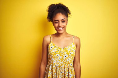 Afro-Amerikalı bir kadın, günlük çiçekli bir elbise giyer, izole edilmiş sarı arka planda durur, kameraya seksi bir ifadeyle bakar, neşeli ve mutlu bir yüzle bakar..