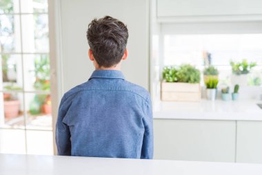 Gündelik gömlekli genç adam beyaz masada oturuyor geriye bakıyor kollarını vücuduna dikiyor.