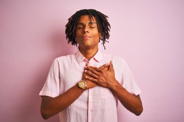 Zarif gömlekli Afro-Amerikan bir adam soyutlanmış pembe arka planda duruyor elleri göğsünde, gözleri kapalı ve yüzünde minnet dolu bir jest ile. Sağlık kavramı.