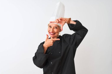 Üniformalı ve şapkalı orta yaşlı aşçı kadın izole edilmiş beyaz arka planda gülümsüyor ve elleri ve parmakları mutlu yüzlü bir çerçeve yapıyor. Yaratıcılık ve fotoğrafçılık kavramı.
