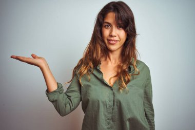 Gri izole arka planda duran yeşil tişörtlü genç ve güzel bir kadın. Neşeli bir şekilde gülümsüyor ve avuç içiyle kameraya bakıyor..