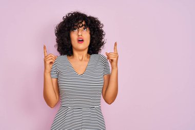 İzole edilmiş pembe arka planda çizgili elbise giyen kıvırcık saçlı genç Arap kadın şaşırmış ve şaşırmış bir şekilde parmaklarıyla işaret ediyor ve kollarını kaldırıyordu..