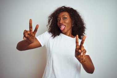 Genç Afro-Amerikalı kadın tişört giyip izole edilmiş beyaz arka planda dikiliyor ve iki elini de zafer işareti yaparken gösteren bir gülümsemeyle gülümsüyor. İki numara..