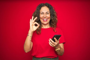 Kırmızı izole edilmiş arka plan üzerinde akıllı telefon kullanan orta yaşlı bir kadın tamam işareti yapıyor, mükemmel bir sembol.