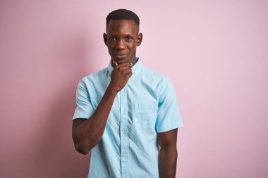 Mavi gömlekli Afro-Amerikalı bir adam pembe arka planda dikilip kollarını kavuşturmuş ve çenesini kaldırarak kameraya gülümsüyor. Olumlu düşünüyorum..