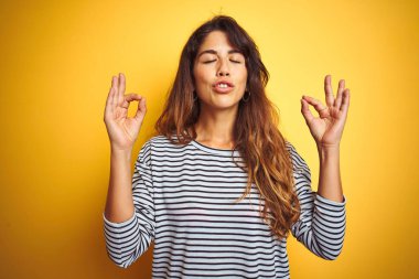Sarı yalıtılmış arka plan üzerinde duran çizgili tişört giyen genç ve güzel bir kadın rahatla ve gözleri kapalı meditasyon yaparken parmaklarınla gülümse. Yoga konsepti.