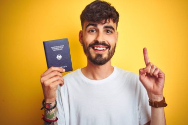 Alman pasaportlu genç adam izole edilmiş sarı arka planda mutlu yüzle işaret eden bir fikir veya soruyla şaşırdı.