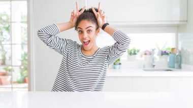 Afro saçlı güzel bir Afro-Amerikalı kadın sıradan çizgili süveter giyiyor komik ve çılgın pozlar veriyor parmakları kafasında tavşan kulağı gibi, neşeli gülüyor