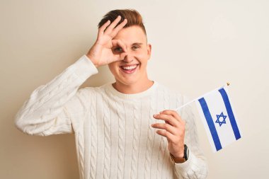 İsrail bayrağını izole edilmiş beyaz arka planda tutan yakışıklı genç adam gülümseyen bir yüzle tamam işareti yaparken parmaklarının arasından bakıyor.