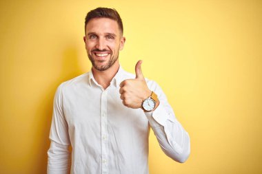 Sarı izole edilmiş arka plan üzerinde zarif beyaz gömlek giyen genç yakışıklı adam mutlu bir el hareketi yapıyor. Kameraya bakıp başarıyı gösteren ifade onaylanıyor.