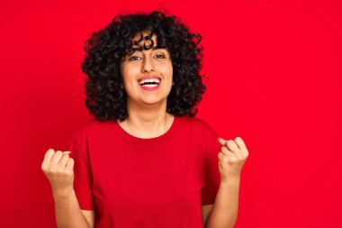 Kıvırcık saçlı genç Arap kadın izole edilmiş kırmızı arka plan üzerine gündelik tişört giyip kolları açık ve açık bir şekilde başarının şaşkınlığını ve şaşkınlığını kutluyor. Kazanan konsept.
