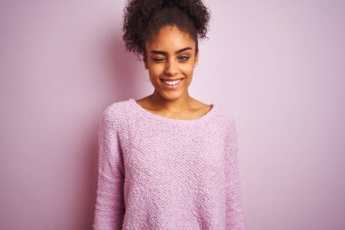 Kış süveteri giyen genç Afrikalı Amerikalı kadın izole edilmiş pembe arka planda dikilip kameraya seksi bir ifadeyle bakıyor, neşeli ve mutlu bir yüzle..