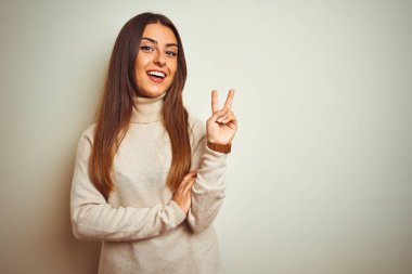 İzole edilmiş beyaz arka planda boğazlı kazak giyen genç ve güzel bir kadın zafer işareti yaparken kameraya gülümsüyor. İki numara..