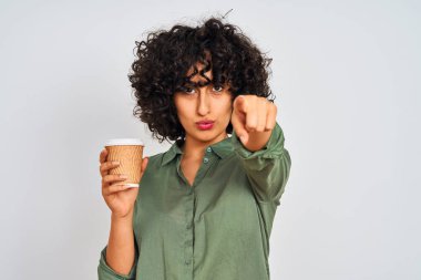 Genç Arap kadın, kıvırcık saçlı, izole edilmiş beyaz arka planda bir fincan kahve içerek kamerayı işaret ediyor ve sen, el işareti, ön taraftan pozitif ve kendinden emin bir hareket.