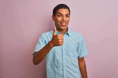 Mavi gömlekli genç, yakışıklı bir Arap, pembe arka planda dikilip mutlu bir el hareketi yapıyor. Kameraya bakıp başarıyı gösteren ifade onaylanıyor.