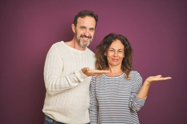 Güzel orta yaşlı bir çift, izole edilmiş mor arka plan üzerine kış süveteri giyip mutlu bir şekilde sunum yapıyor ve avuç içi kameraya bakıyor..
