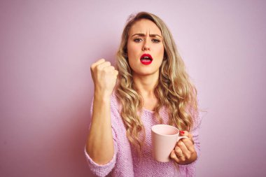 Genç, güzel bir kadın pembe izole edilmiş arka planda bir fincan kahve içerken sinirlenmiş ve sinirlenmiş bir şekilde bağırıyor, çıldırmış ve el kaldırarak bağırıyor.