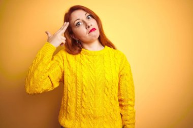 Kışlık kazak giyen güzel bir kızıl kadın izole edilmiş sarı arka planda duruyor. Kendini vuruyor ve öldürüyor. Silah gibi el ve parmakları işaret ediyor..