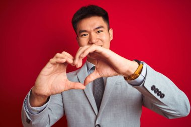 Asyalı Çinli işadamı gri ceketli ve kravatlı, izole edilmiş kırmızı arka planda duruyor. Aşkla gülümsüyor, elleriyle kalp sembolü ve şekli gösteriyor. Romantik konsept.