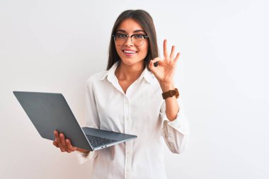 Gözlük takan güzel bir iş kadını izole edilmiş beyaz arka plan üzerinde dizüstü bilgisayar kullanıyor tamam işareti yapıyor, mükemmel bir sembol.