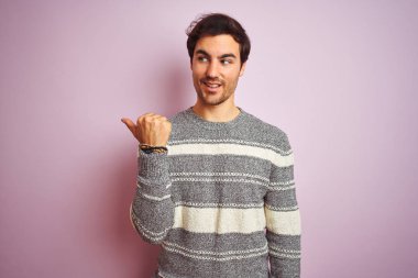 Sıradan çizgili süveter giyen genç, pembe arka planda duran mutlu yüzüyle gülümseyen ve baş parmağıyla yan tarafı gösteren yakışıklı bir adam..