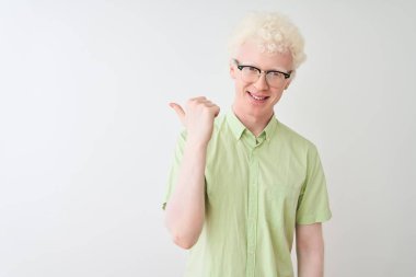Yeşil tişörtlü ve gözlüklü genç albino adam izole edilmiş beyaz arka planda gülümsüyor ve başparmağıyla yana bakıyor..