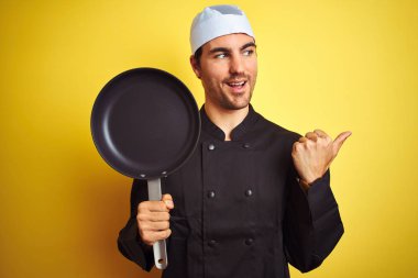 Üniformalı ve şapkalı genç aşçı, izole edilmiş sarı arka planda tavayı tutuyor ve gülümseyen bir gülümsemeyle baş parmağıyla onu işaret ediyor.