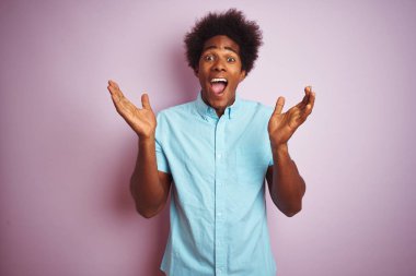 Afro saçlı, mavi tişörtlü genç bir adam, izole edilmiş pembe arka plan üzerinde durmuş çılgın ve başarıyı kutlarken kollarını kaldırmış ve gözleri açık bir şekilde heyecanla haykırıyordu. Kazanan konsept
