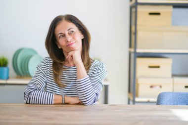 Orta yaşlı bir kadın evinde oturmuş kameraya kendinden emin bir şekilde bakıyor kollarını kavuşturmuş ve çenesini kaldırmış gülümsüyor. Olumlu düşünüyorum..