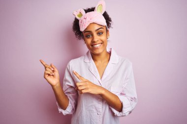 Genç Afro-Amerikan kadın pijama giyiyor ve maske takıyor pembe arka planda gülümsüyor ve kameraya bakıyor iki eliyle ve parmaklarıyla yandan işaret ediyor..