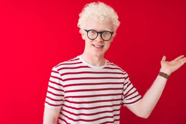 Genç albino sarışın adam çizgili tişört ve gözlüklerle izole edilmiş kırmızı arka plan üzerinde gülümseyen ve eli kameraya bakarak elini gösteren..
