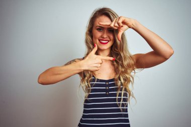 İzole edilmiş beyaz arka plan üzerinde duran çizgili tişört giyen genç ve güzel bir kadın gülümsüyor ve elleri ve parmakları mutlu yüzlü bir çerçeve yapıyor. Yaratıcılık ve fotoğrafçılık kavramı.