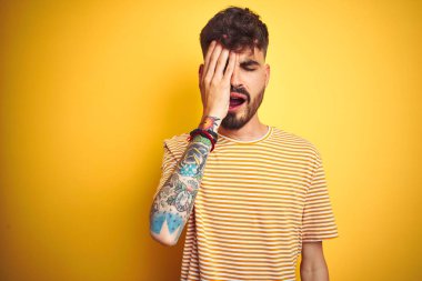 İzole edilmiş sarı arka plan üzerinde duran çizgili tişörtlü genç adam esnemekten yoruldu. Yüzünün yarısını, gözünü ve ağzını eliyle kapattı. Yüzüm acıyor..
