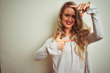 Genç ve güzel bir kadın izole edilmiş beyaz arka planda duruyor gülümsüyor ve elleri ve parmakları gülen yüzlü bir çerçeve yapıyor. Yaratıcılık ve fotoğrafçılık kavramı.