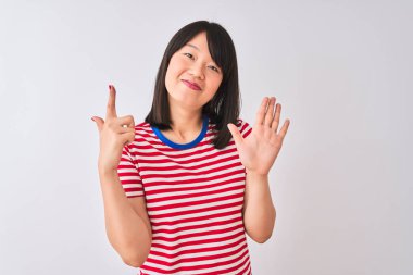 İzole edilmiş beyaz arka plan üzerinde kırmızı çizgili tişört giyen genç ve güzel Çinli kadın yedinci parmaklarını gösterirken kendinden emin ve mutlu bir şekilde gülümsüyor..