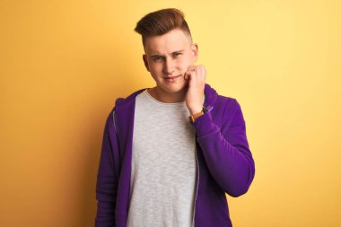 Mor eşofman üstü giyen genç bir adam izole edilmiş sarı arka planda durup diş ağrısı ya da diş ağrısı yüzünden ağız ve acı dolu bir ifadeyle dokunuyor. Dişçi konsepti.